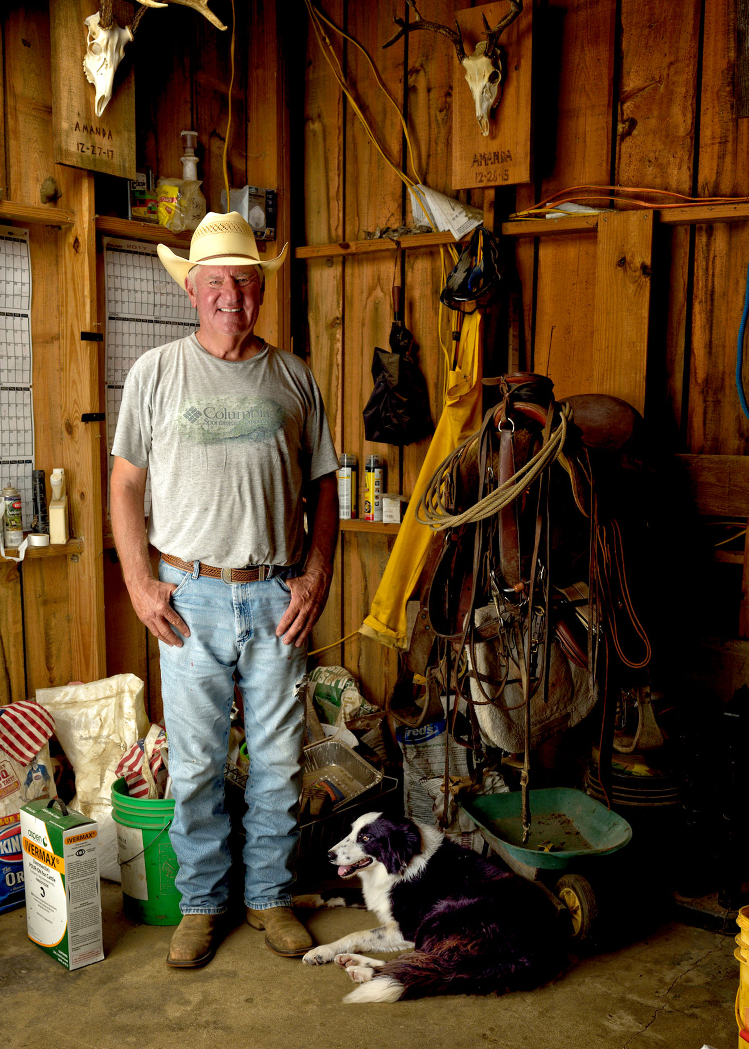 A man wearing a cowboy hat stands in a barn with horse riding equipment behind him and a brown and white dog beside him. 