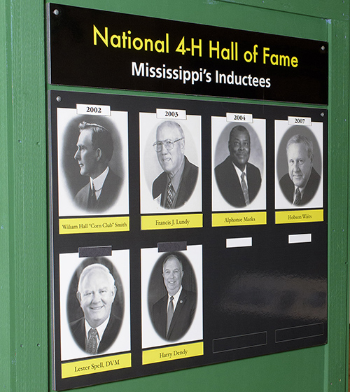 A green board with the words “National 4-H Hall of Fame Mississippi’s Inductees” and pictures of six men stands in a building with a Mississippi map in the background.