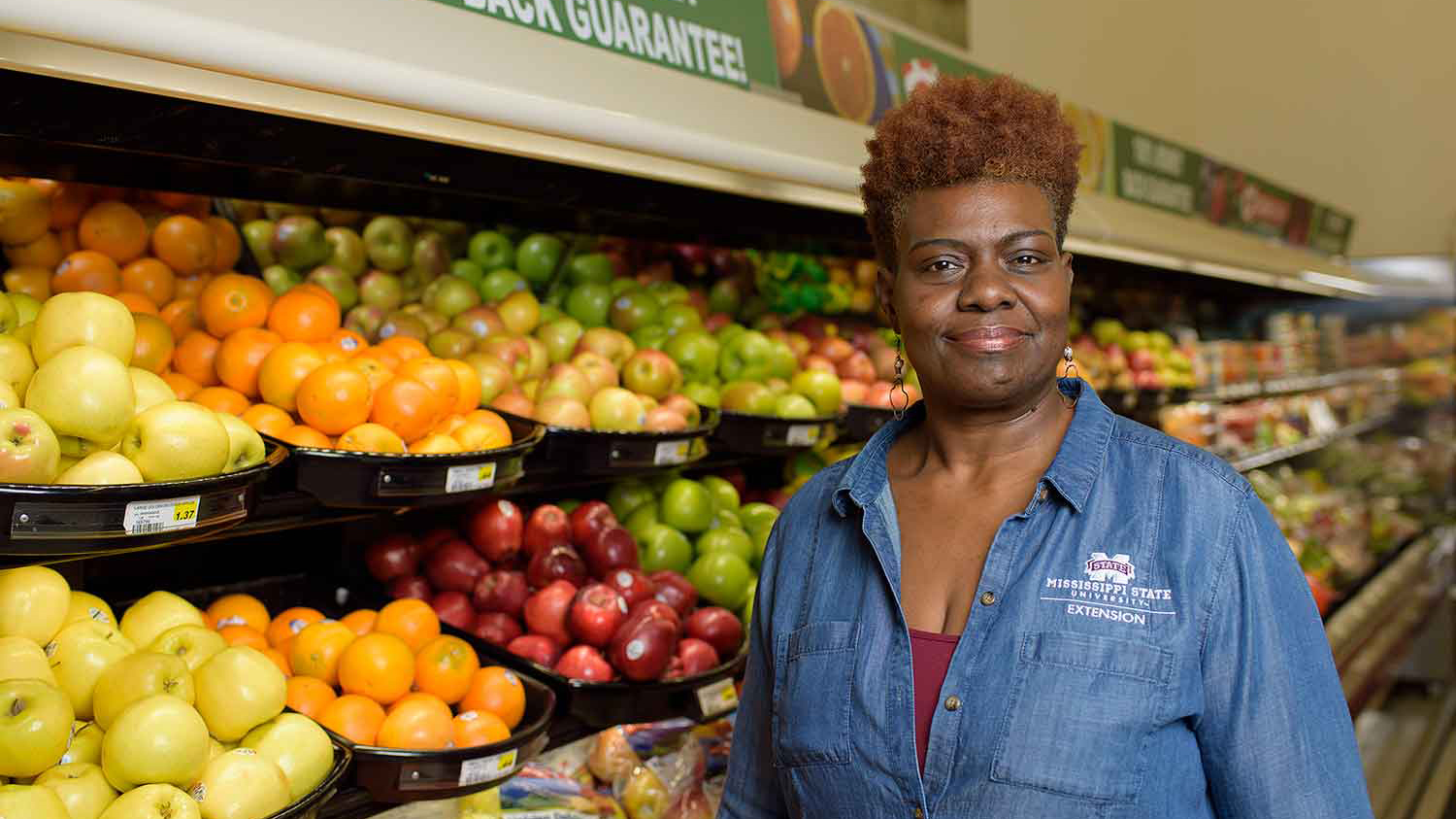 A woman, wearing a denim shirt, smiles in front of a fruit display at a grocery store.