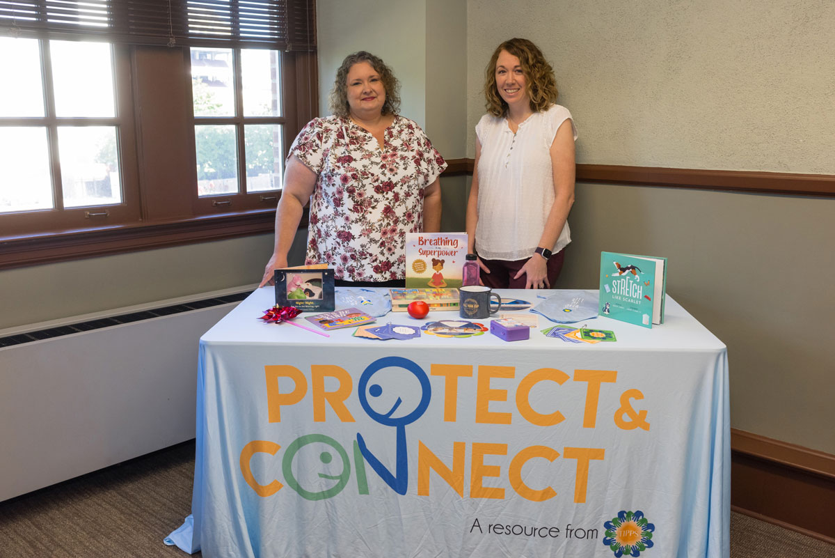 Two women stand behind a table with a cloth that reads “Protect & Connect.”