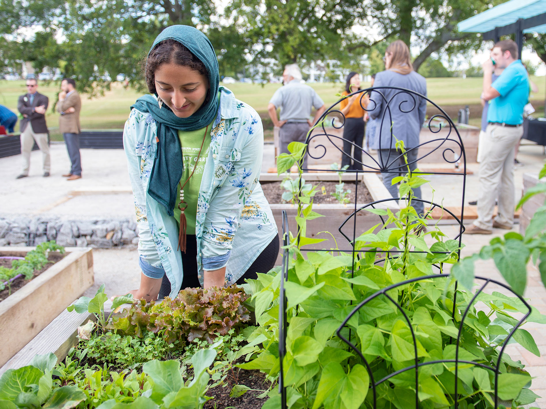 A woman with a teal scarf wrapped around her head leans toward a gardening box filled with bright green, leafy plants. Several other people stand conversing in the background.