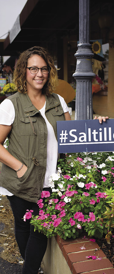 A woman stands on Main Street, beside a street lamp, holding a #Saltillo sign.