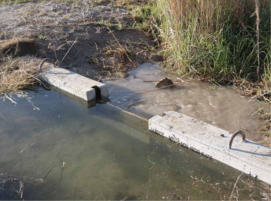 A wier, or concrete structure placed across the width of a drainage ditch to control the flow of water off of an agricultural field. There is a small space between two structures for water to flow through.
