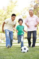 A grandfather, father, and young son play soccer together in a park.