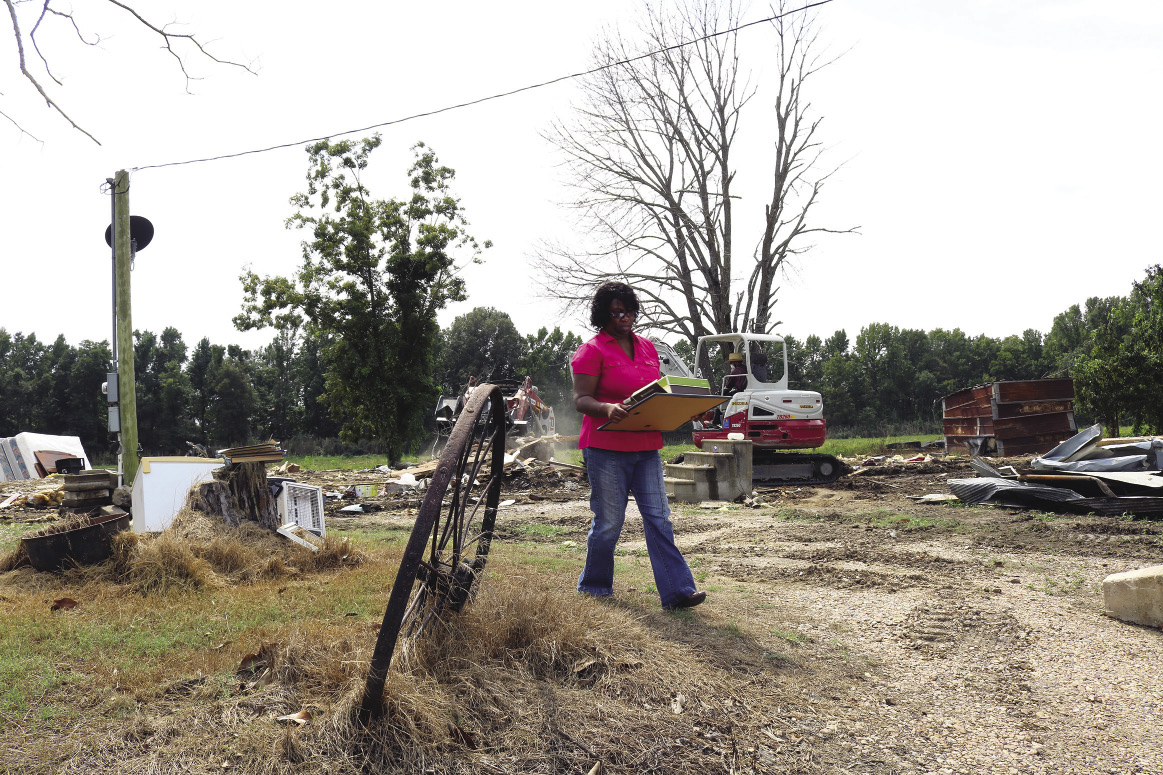 A woman carries some of her belongings at the site of a destroyed house. A piece of heavy equipment demolishes the house in the background.