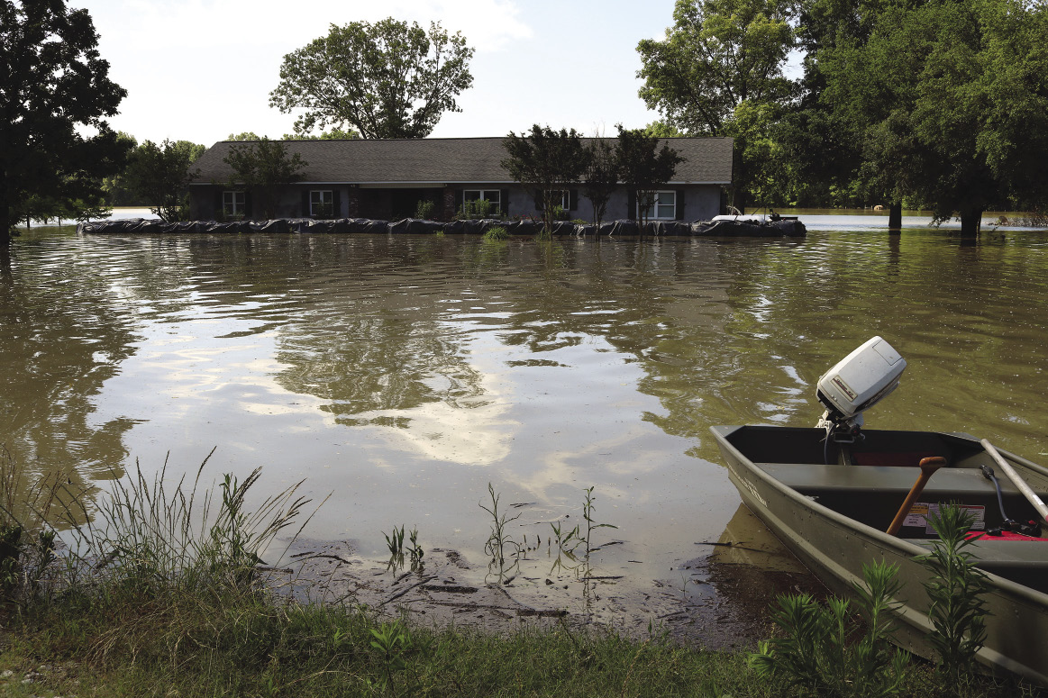 A house has a sand-bag levee keeping out the surrounding flood water. Across the water on dry land, a small boat is moored.