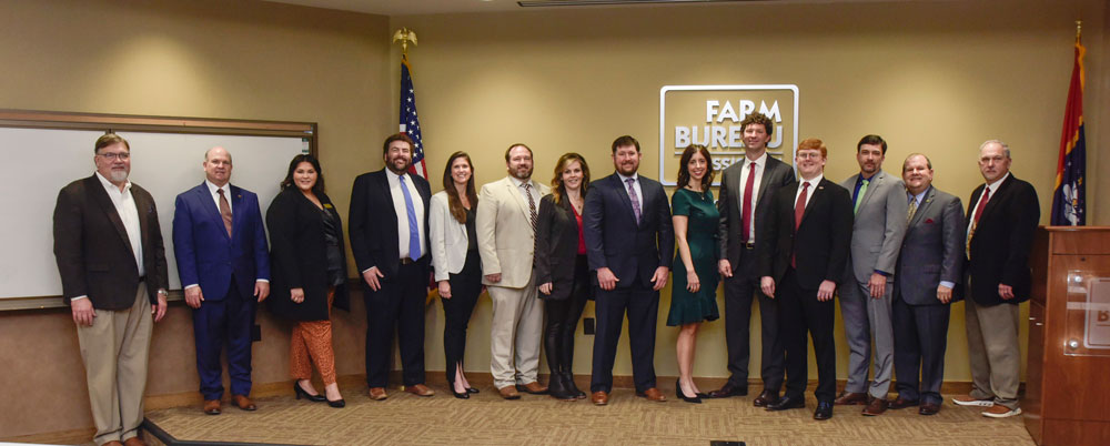 Fourteen smiling men and women stand in front of the Mississippi Farm Bureau seal.