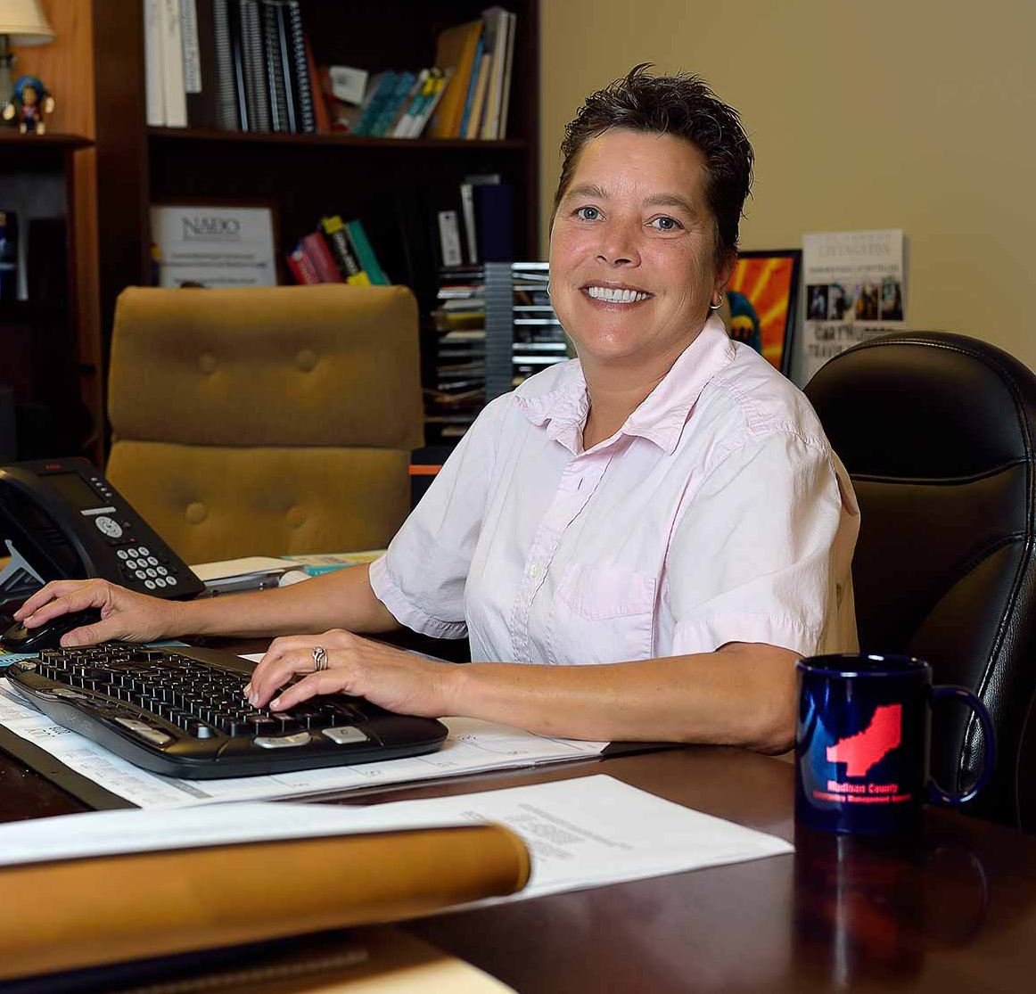 A woman with short hair and a collared, light pink shirt sits at her desk smiling. Her desk has papers and notebooks on it, along with a coffee mug, keyboard, and office phone.