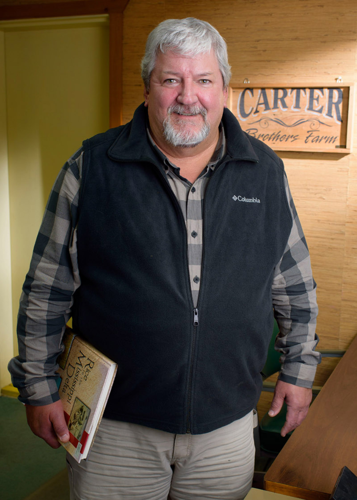 A gray-haired man with a goatee wears a navy blue vest over a blue-checkered, long-sleeved polo and tan slacks. He stands in front of a “Carter Brothers Farm” sign and beside a desk with a rolodex as sun filters across the background.