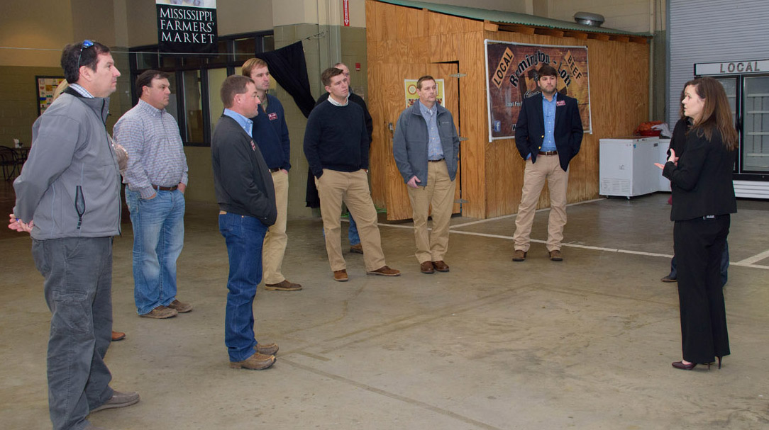 7 men, standing, listen to a woman wearing black and standing in front of a “Local Beef” refrigerator