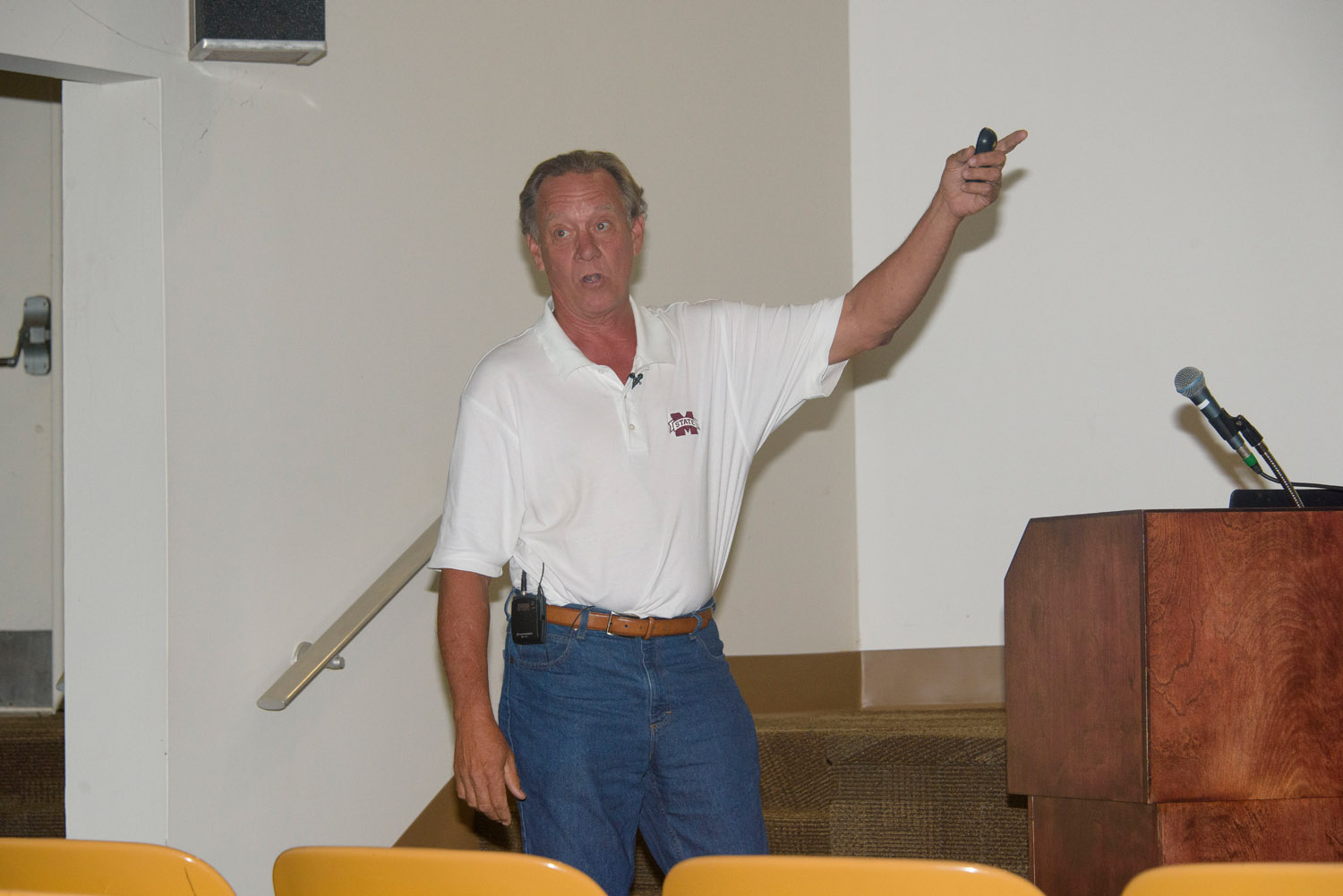 A man wears a white polo shirt with a Mississippi State University emblem on the right breast pocket, and his arm is lifted and pointing right.