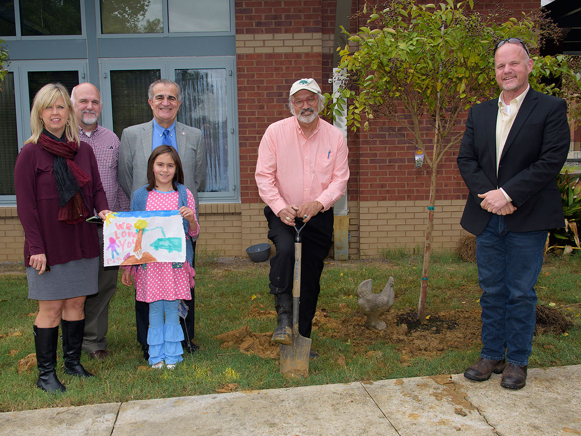 Several people stand outside of a brick building and next to a newly planted tree. 