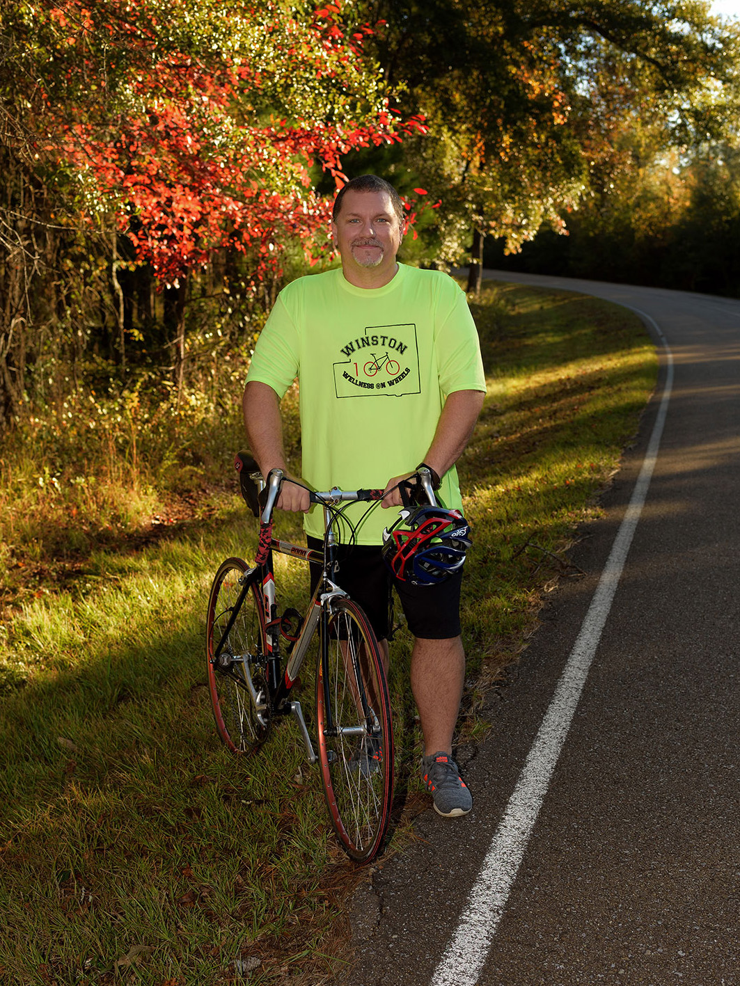 A man wearing a fluorescent green shirt stands holding the handlbars of a black and red bike on the side of a road.
