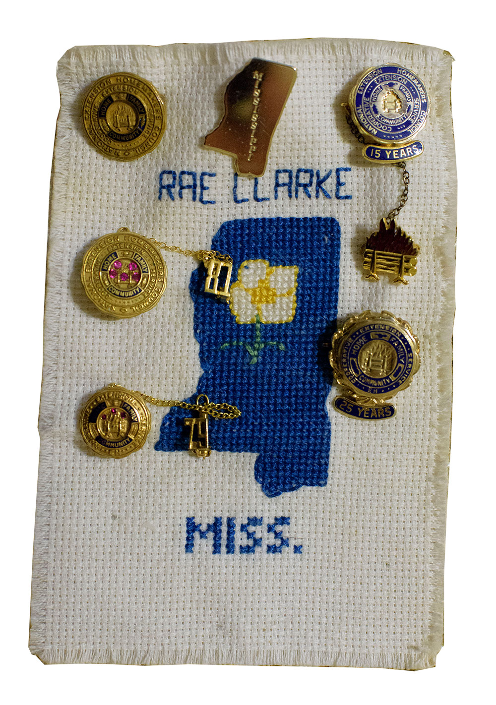 A rectangular piece of white fabric has a blue outline of the state of Mississippi and the words "Rae Clarke" and "Miss." The fabric is decorated with several gold pins.