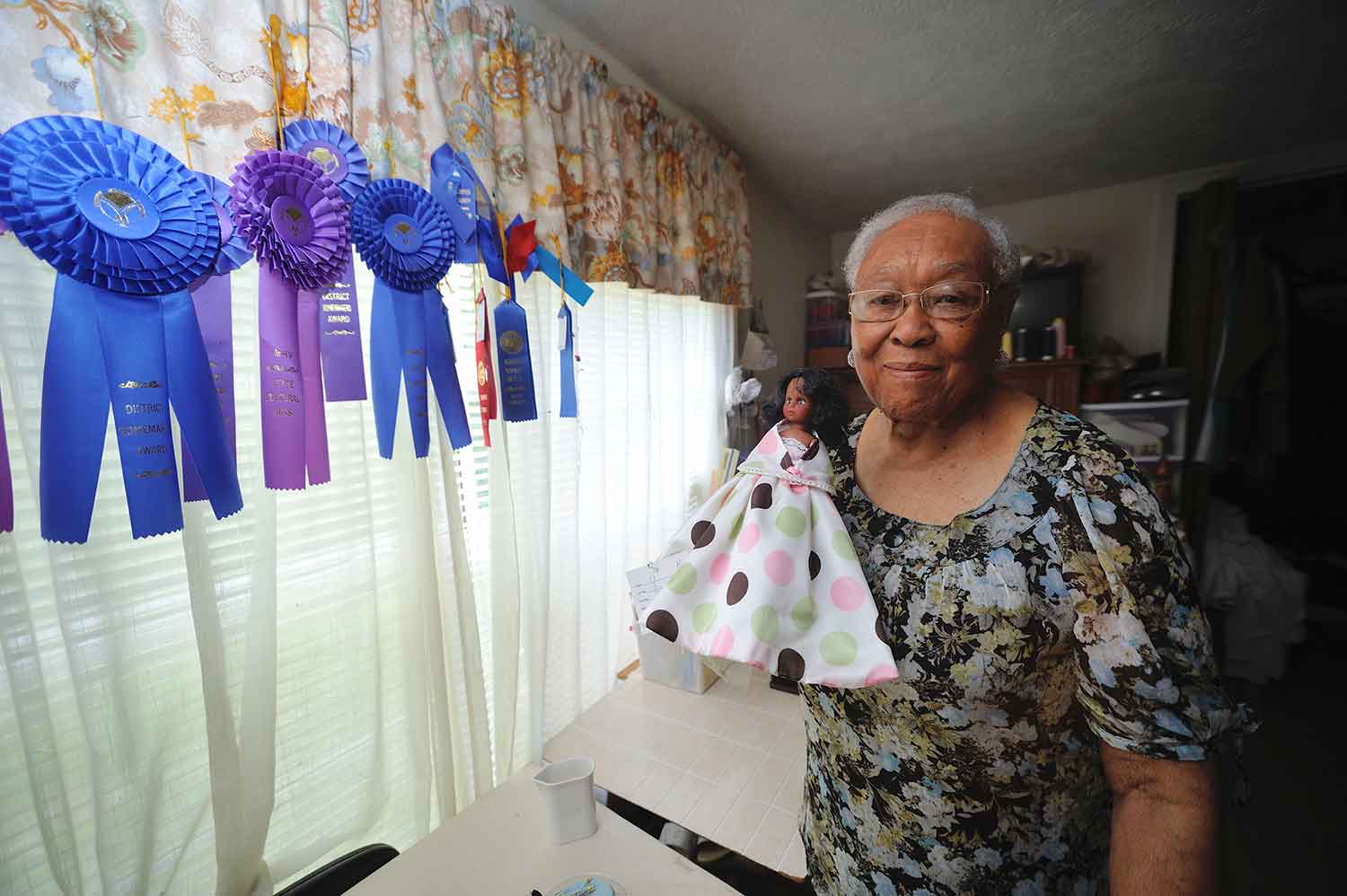 An woman with short grey hair stands to the right of a windowsill with blue and purple ribbons hanging from a multicolored curtain. The woman holds a doll wearing a white dress with light green, light pink, and dark brown polka dots.