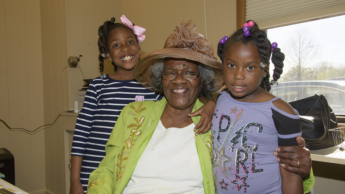 A young girl wearing a purple shirt, a young girl wearing a striped shirt, and an older woman wearing a large brown hat and a bright green jacket smile at the camera.