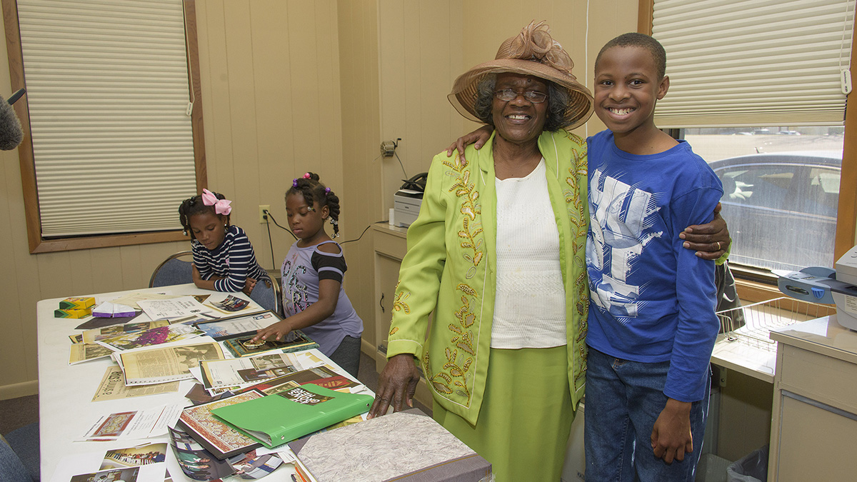 A woman in a large brown hat and a bright green jacket smiles next to a young boy wearing a dark blue shirt.