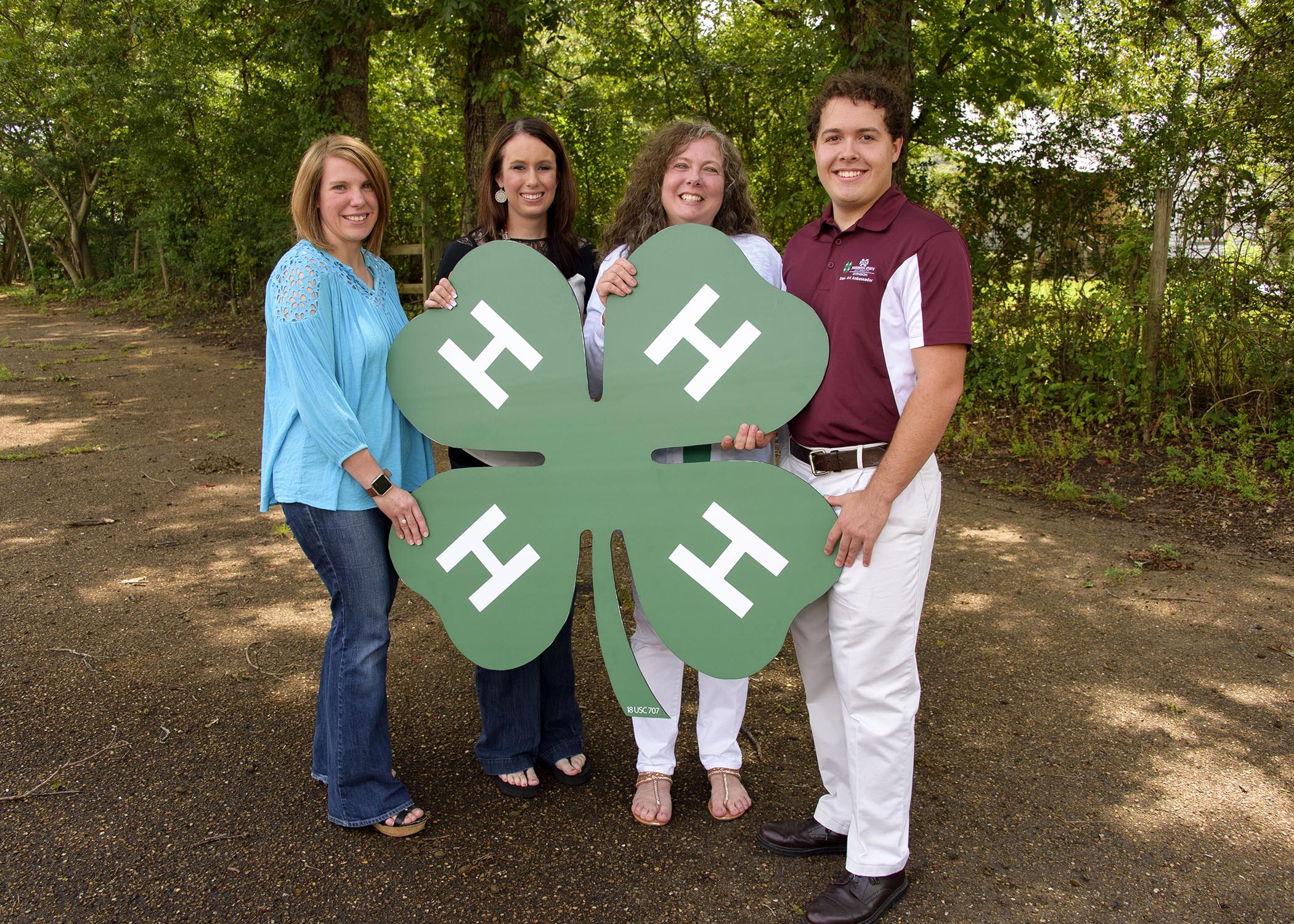 Three females and one male smile as they hold a wooden cutout of the 4-H logo.