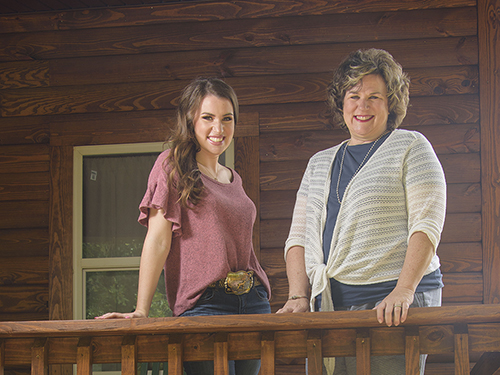 On the left, a young woman with dark brown hair, blue jeans, and pink shirt tucked in, showing a belt with a large golden belt buckle, stands beside an older woman with short brown hair, a blue shirt, and a white cardigan stands.  