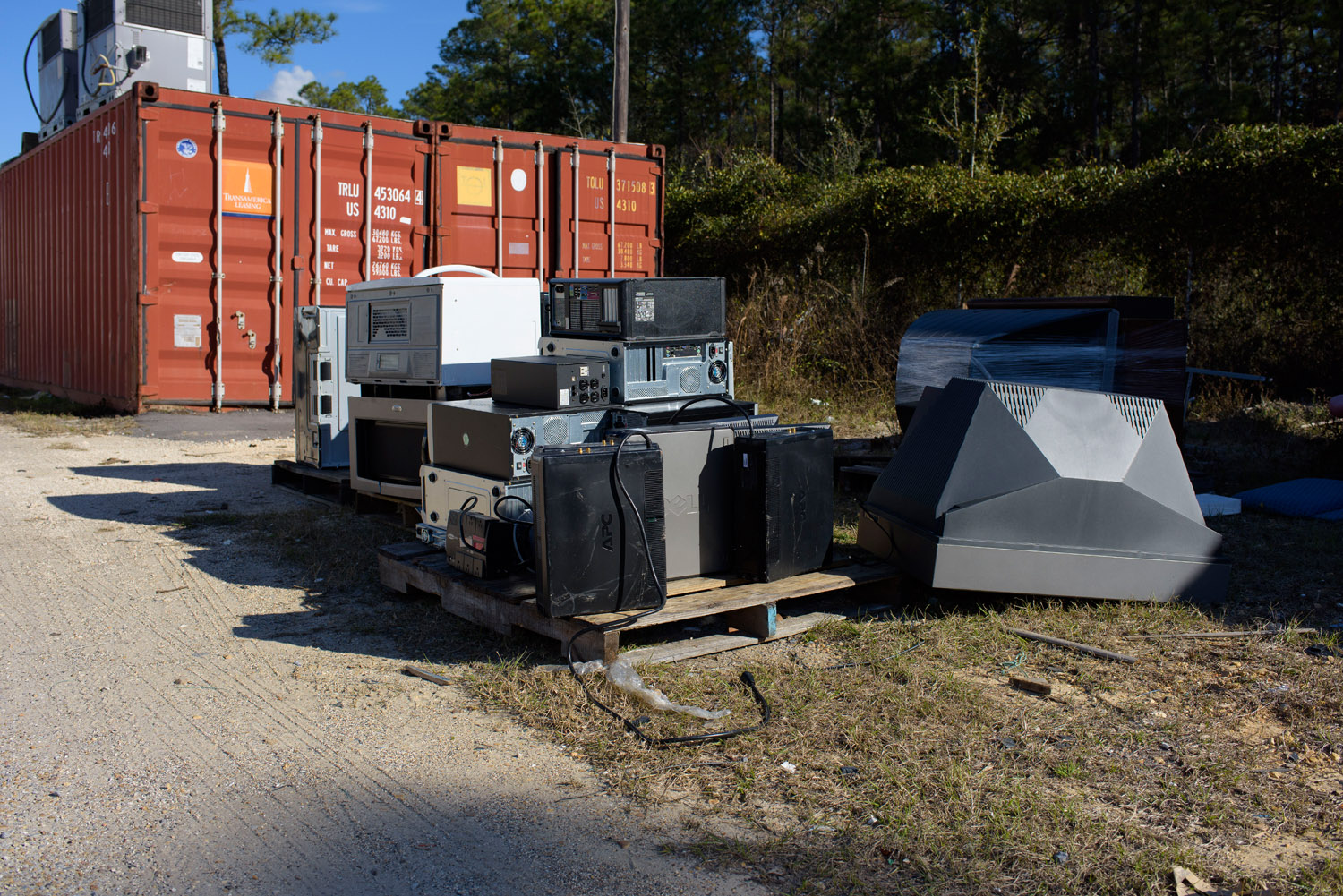 In front of a railroad car on the sand, an overturned tube television, right, shines beside a pallet loaded with old VCRs, CPUs, and microwaves.