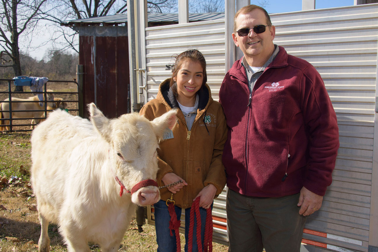 A female teen in a brown 4-H jacket, and jeans stands smiling between a man wearing sunglasses and a maroon pullover and a white cow.