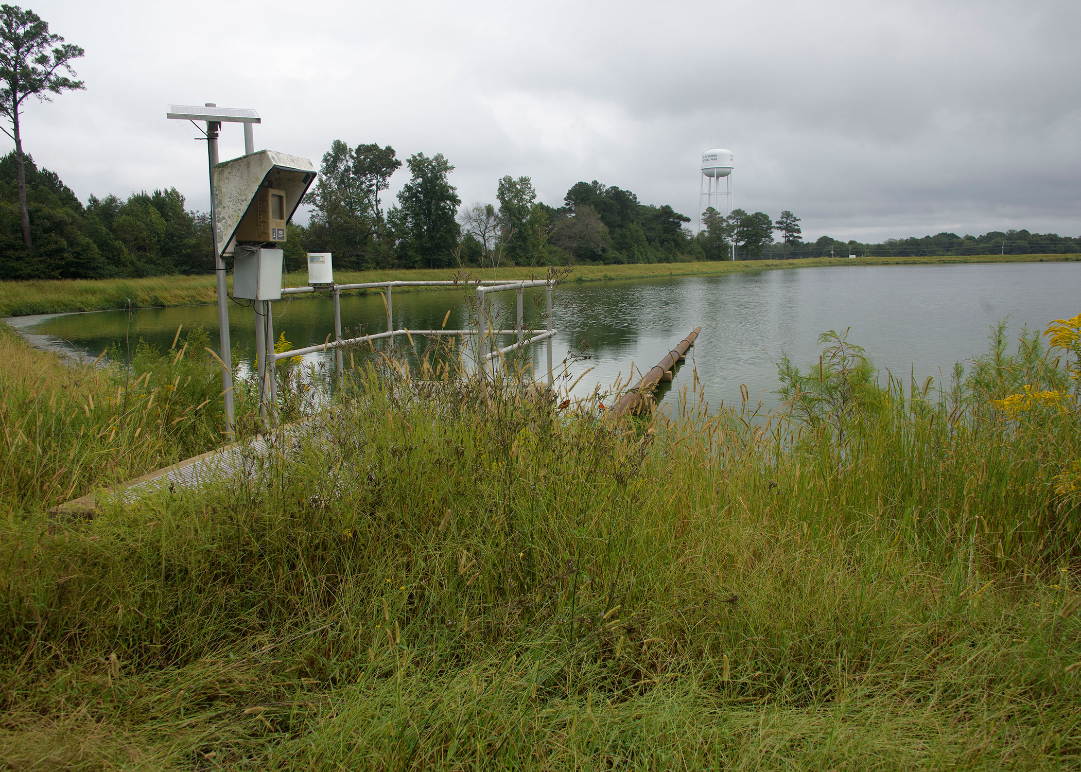 A metal structure stands in tall, green grass on the edge of a pond, where a large pipe reaches into the water from the metal structure. 