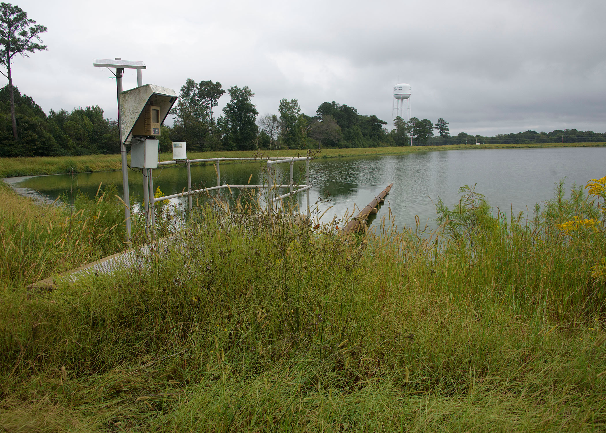 A metal structure stands in tall, green grass on the edge of a pond, where a large pipe reaches into the water from the metal structure.