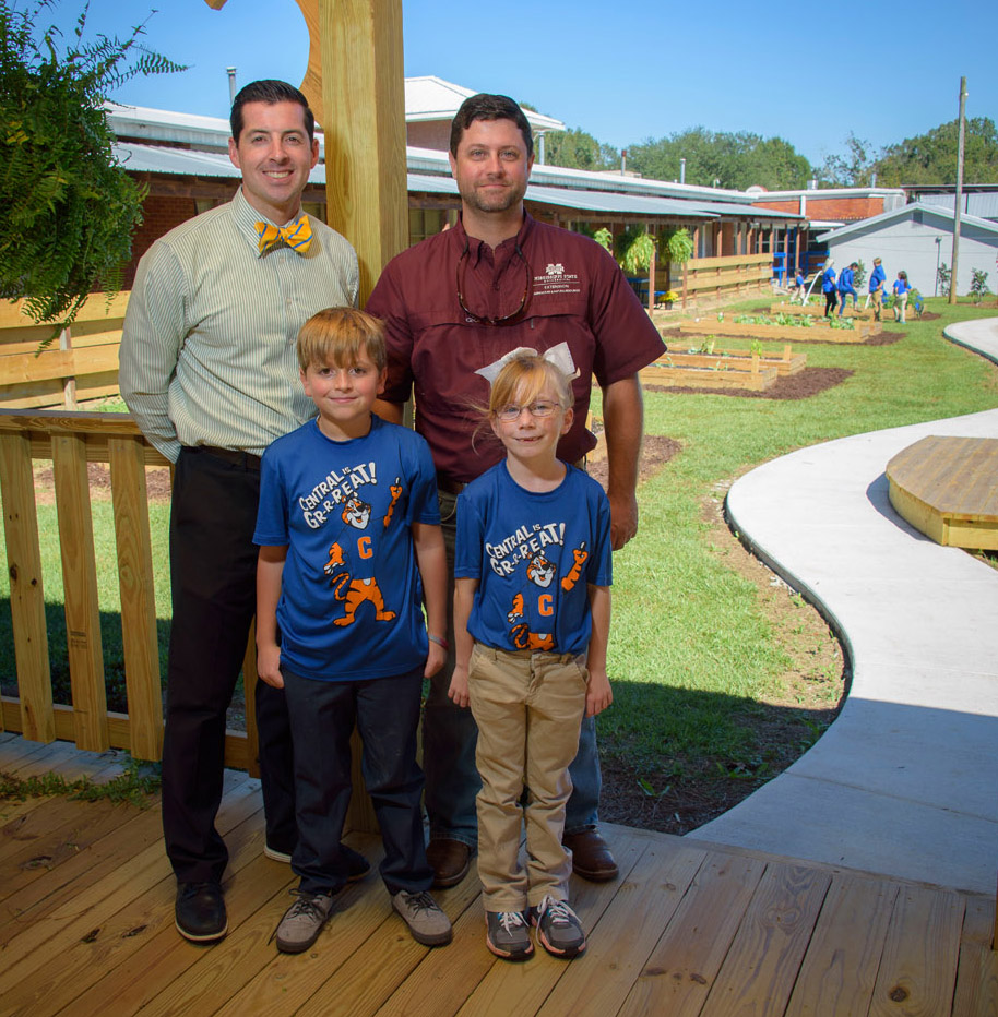 Two men, one on the left in a white & blue striped polo and one on the right with a maroon polo, stand behind a male and female child, both blonde and wearing T-shirts with “Central is Gr-r-reat!” and a tiger printed on them. They all stand on a wooden patio in front of a small farm site.