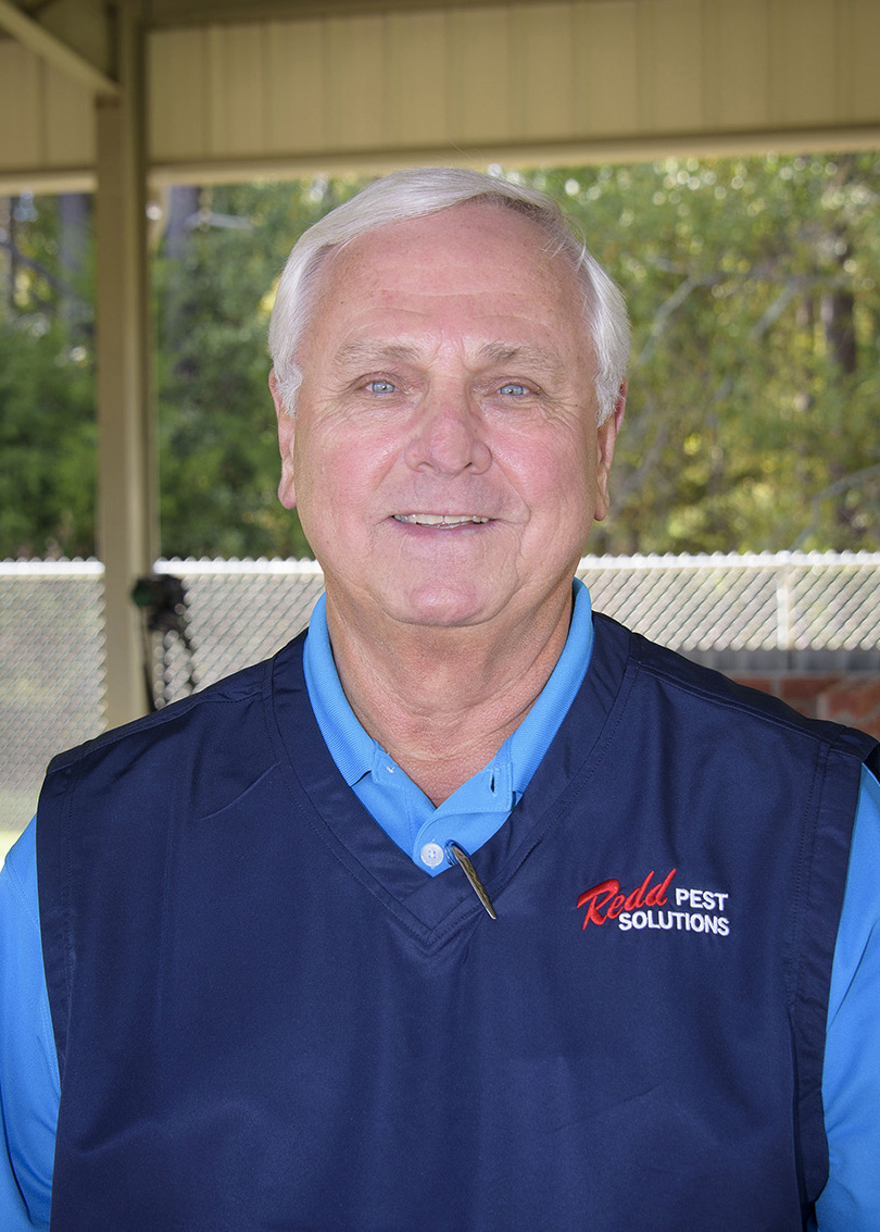 A white-haired man wearing a dark blue collared shirt and a black vest stands smiling at the camera.