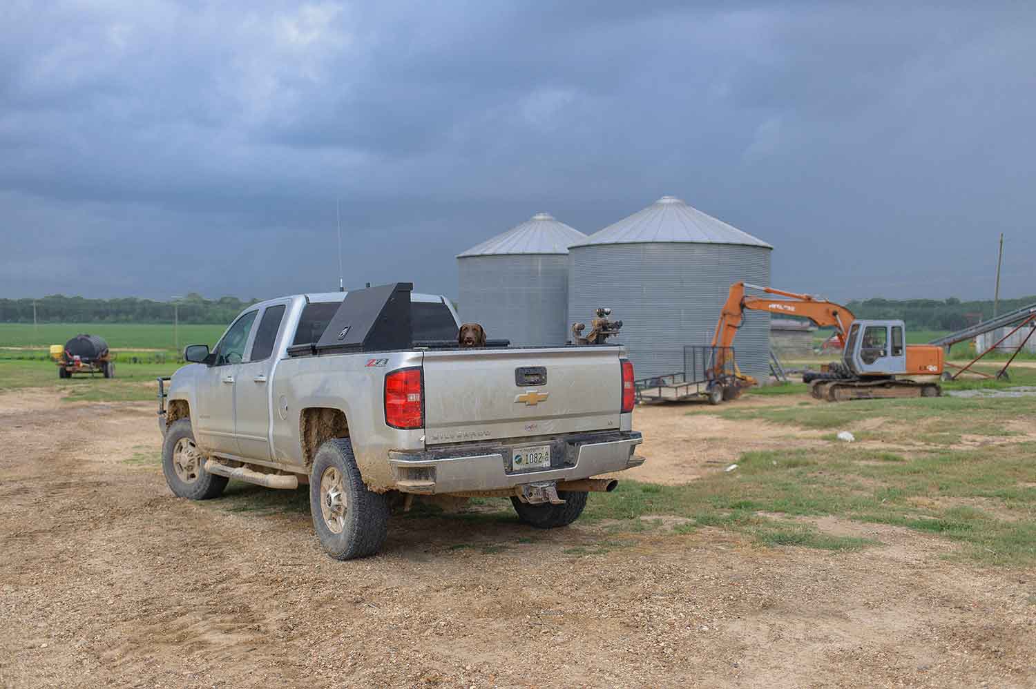 A silver truck with a brown dog peeking its head out of the bed is parked on a dirt lot with farms in the background. Two large, round, metal buildings are just past the truck, and an orange bulldozer is parked in front of one of the metal buildings