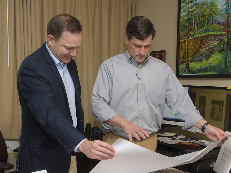 Two men stand behind a desk while looking at and holding a large piece of paper.