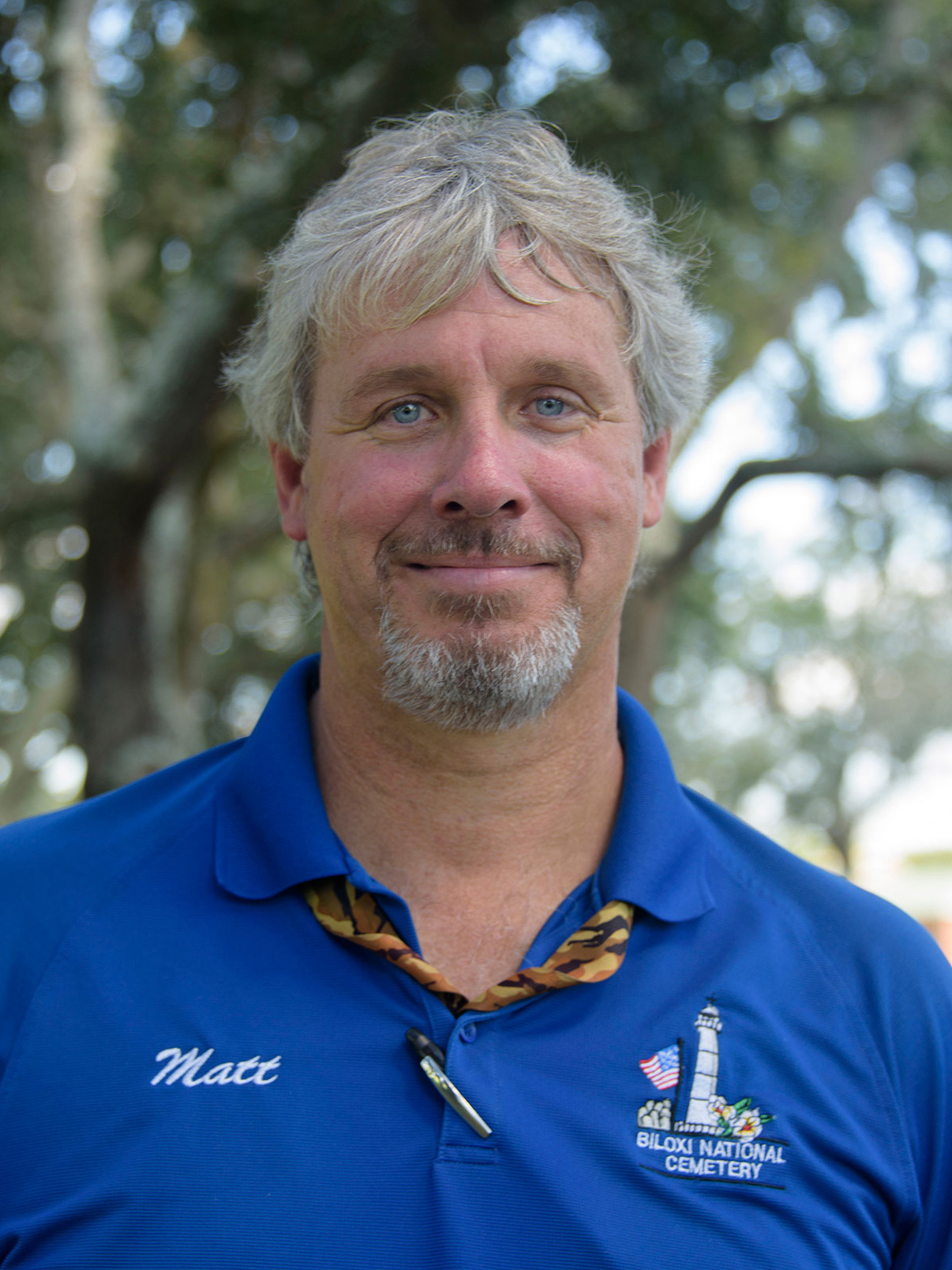 A grey-haired man wearing a blue collared shirt smiles at the camera.