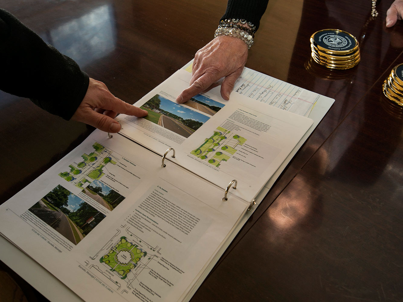 A binder with plans for the city is open on a table, and tow hands are pointing at a picture of a road in the binder.