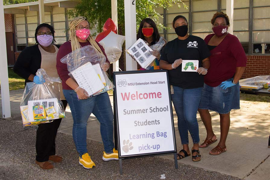 Five women wearing maks and holding plastic bags filled with learning material stand behind a sign that says, “Welcome Summer School Students Learning Bag pick-up.”