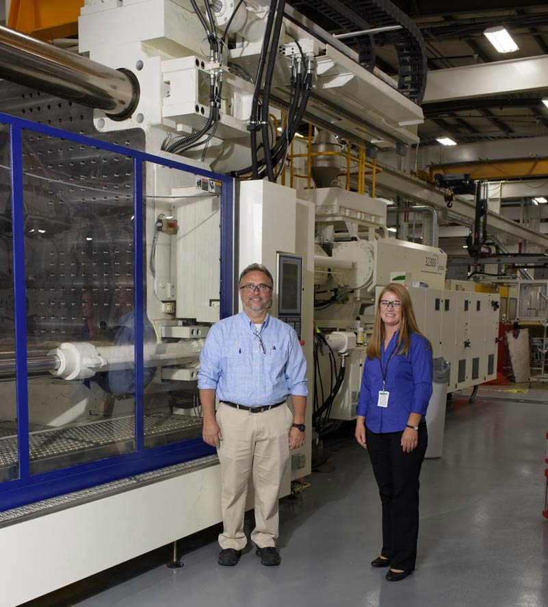 A man and woman wearing safety goggles stand in front a large piece of machinery in a warehouse.