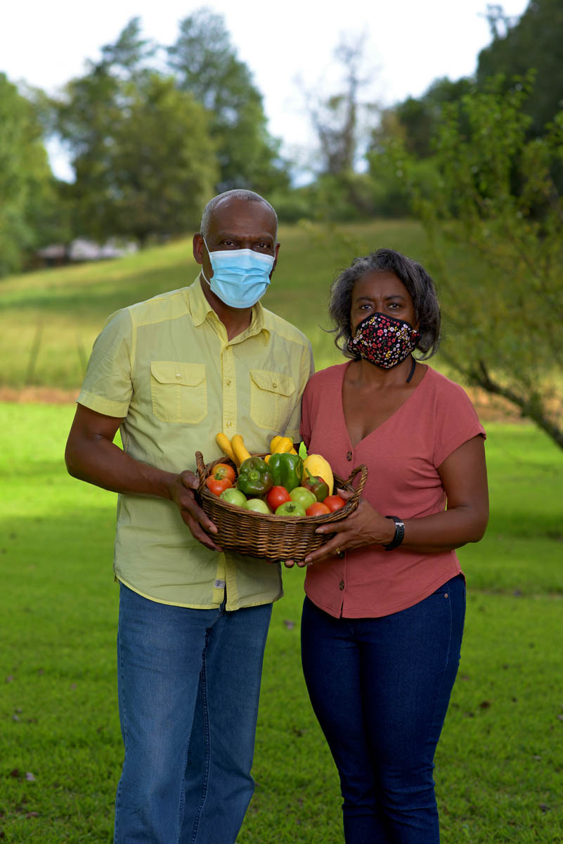A man wearing a yellow shirt and blue mask and a woman wearing a pink shirt and floral mask stand next to each other, both holding a basket of fresh vegetables including tomatoes, bell peppers, and squash.