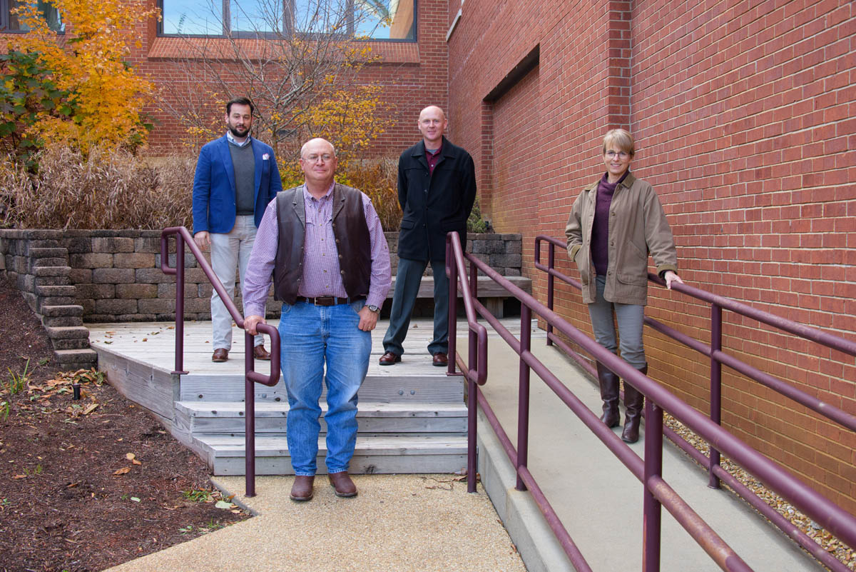 Three men and one woman standing, spaced out in front of a red brick building.