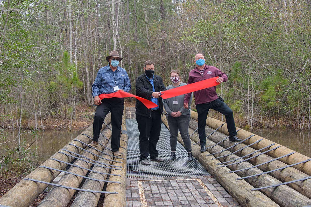 Four people stand behind a recently cut large red ribbon on a new bridge over a body of water. 