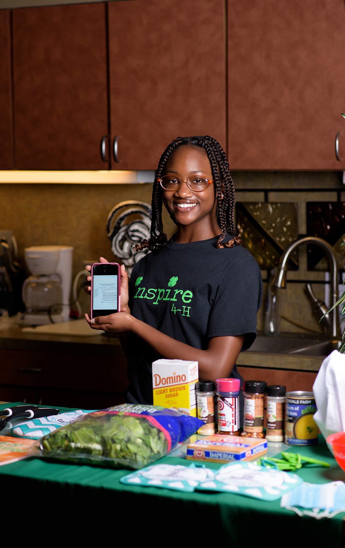 A smiling teenage girl stands behind a table covered in ingredients for a recipe displayed on a phone screen she holds in front of her. 