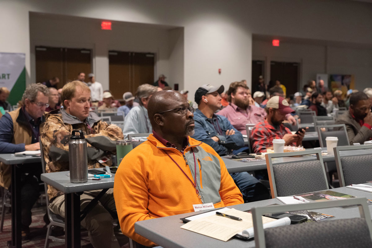 : Tables of people look forward at a speaker (not pictured). A man wearing an orange jacket is in the foreground.