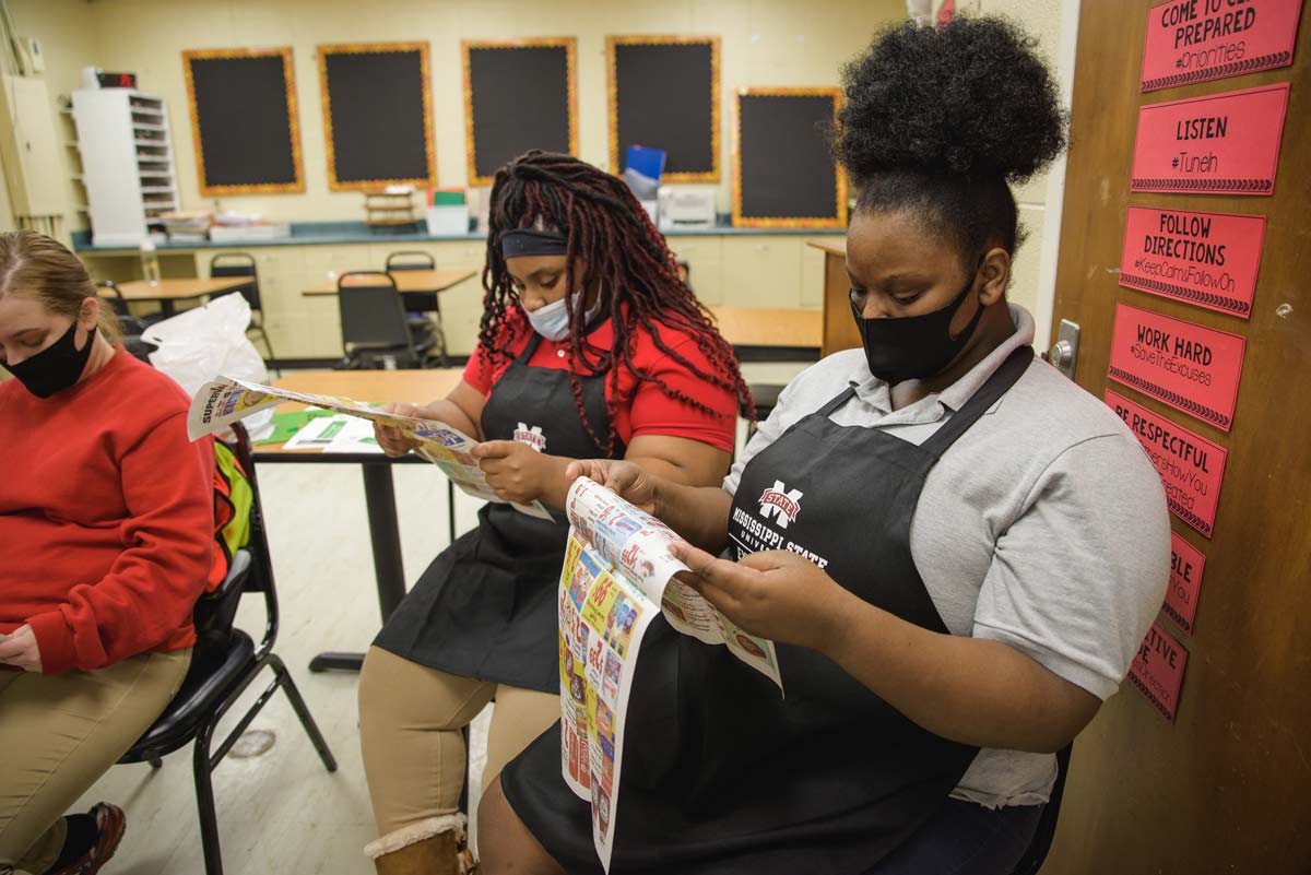 Two young women, wearing aprons and facemasks, look at grocer circulars.