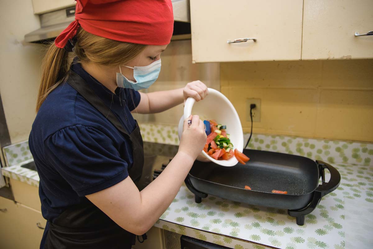 A female teen wearing a chef’s hat transfers vegetables into a cooker.