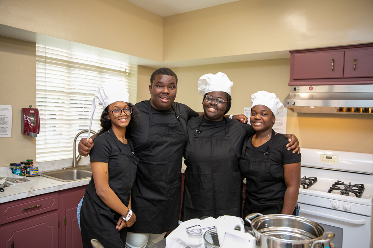 Four teenagers wearing black aprons and white chef hats smile for a group photo.
