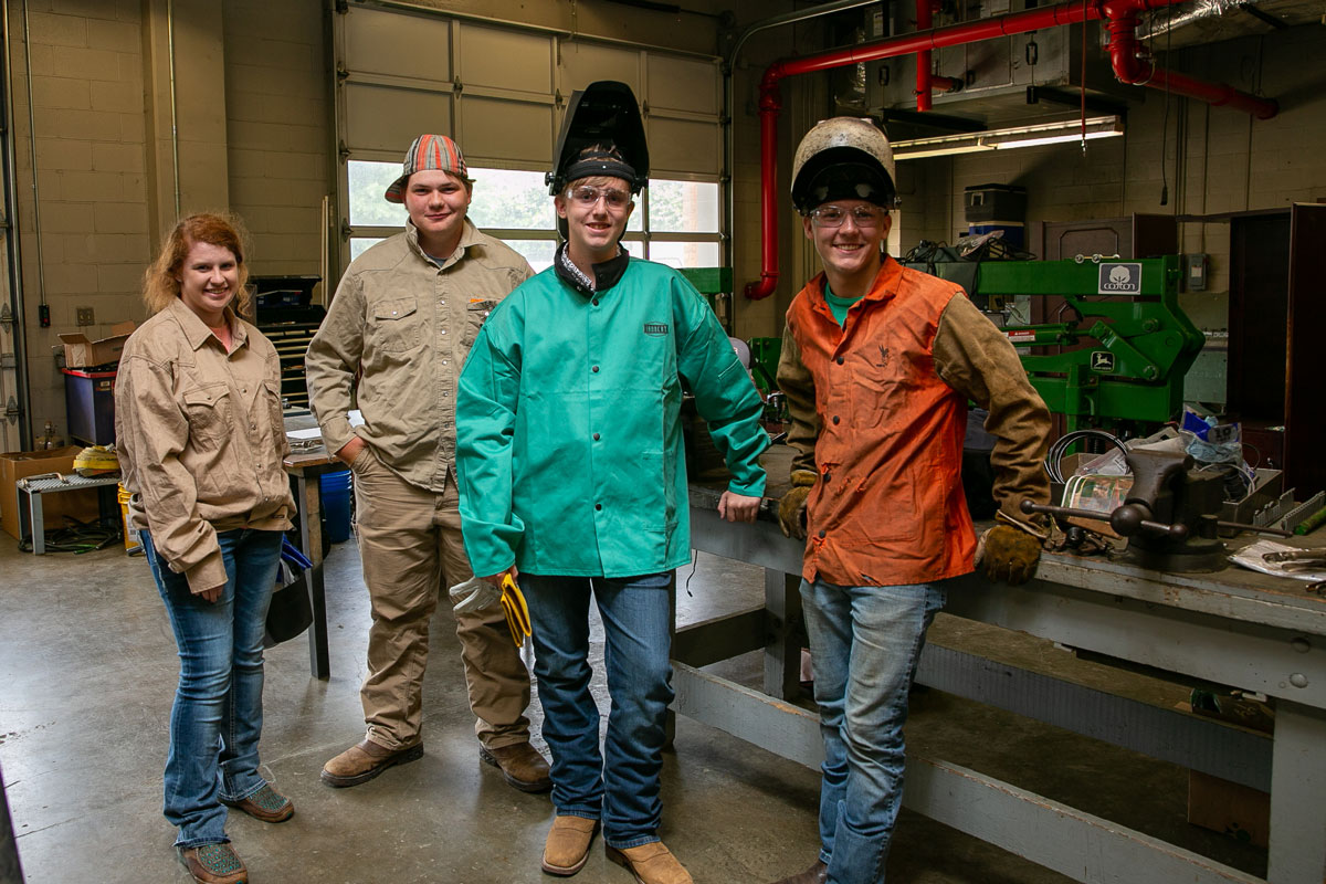 Three young men and one young woman smile for a group photo. Two of the boys wear welding helmets.
