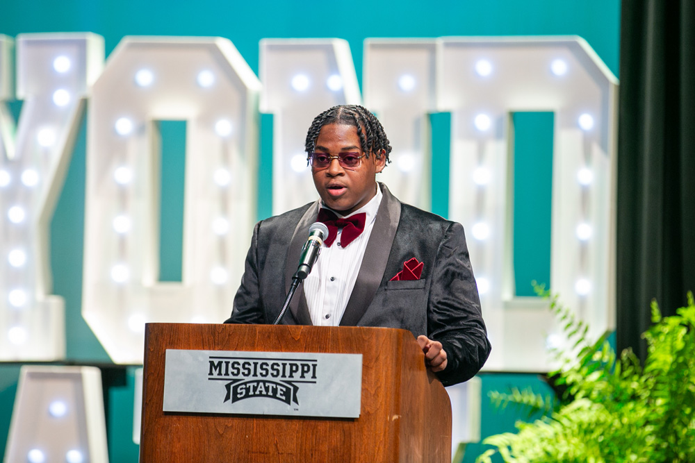 A teen dressed in a suit speaking at a podium.