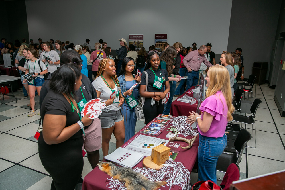 Three teens talk to a person at a maroon display table featuring handouts and themed decorative items. Two other teens observe.