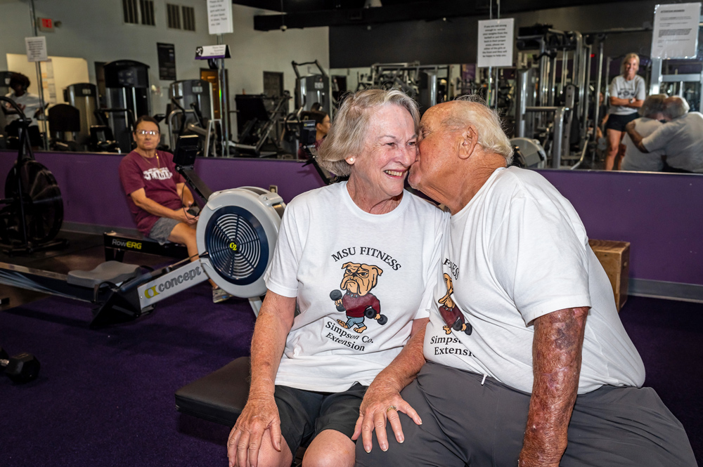 A couple sitting together in a gym; one person gently rests their face against their partner’s face.