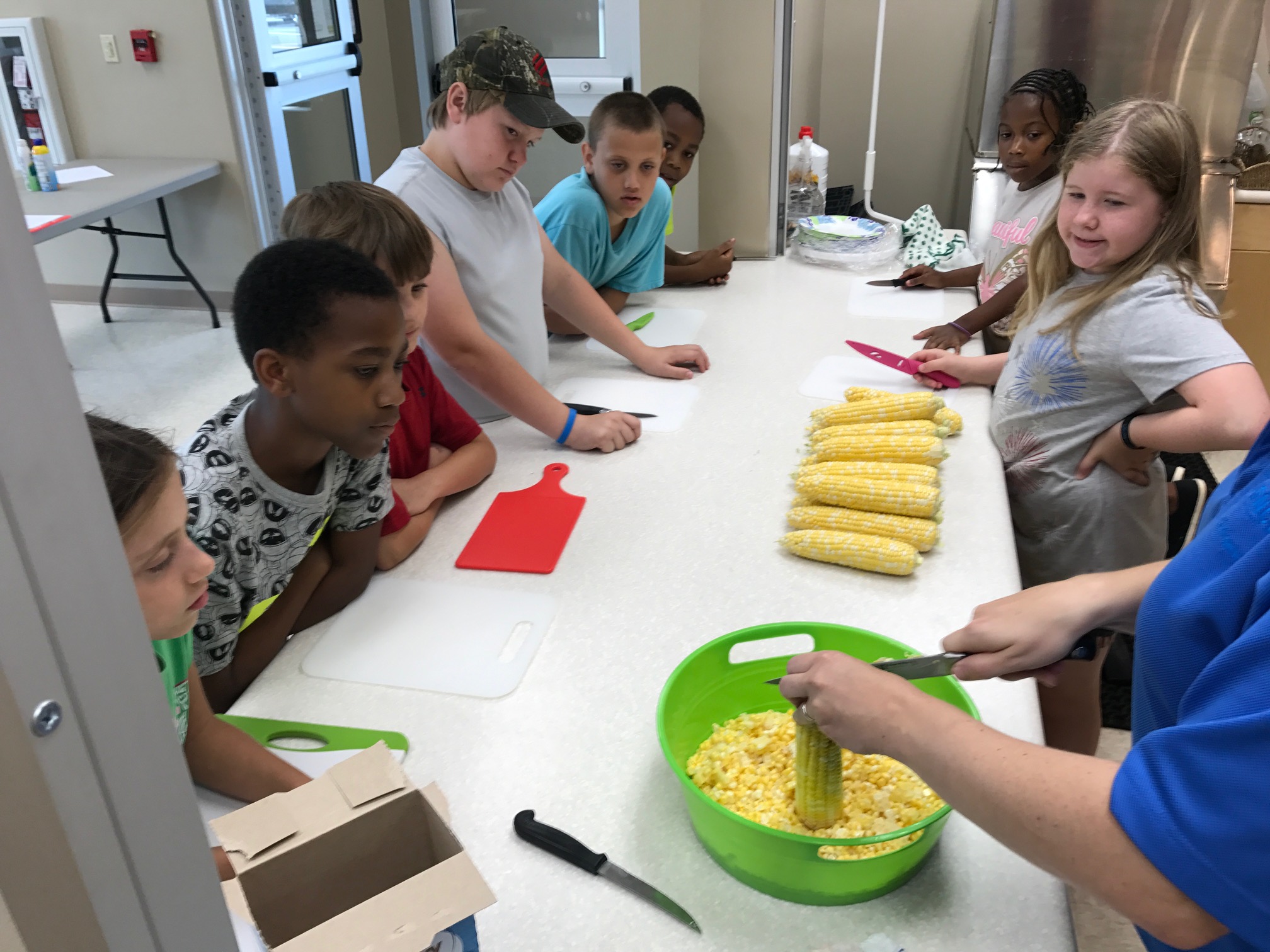 Children watch as an adult cuts corn kernels from the ear.