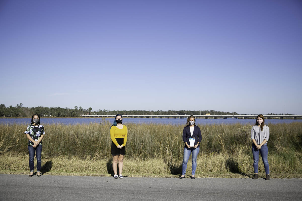 Four women stand spaced out in front of tall grass surrounding bright blue water.