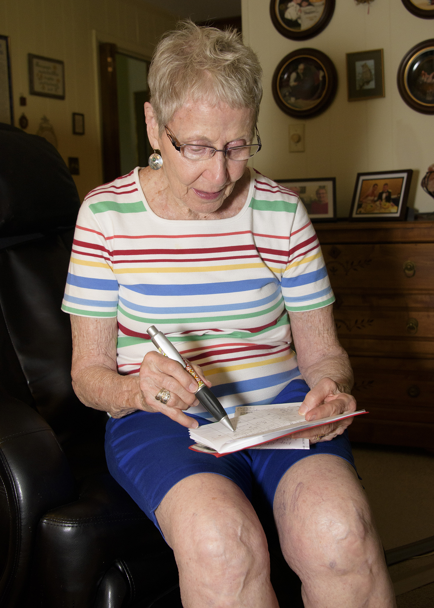 A gray-haired woman in blue shorts and a multi-colored striped shirt holds a pen over a pocket calendar to check appointments.